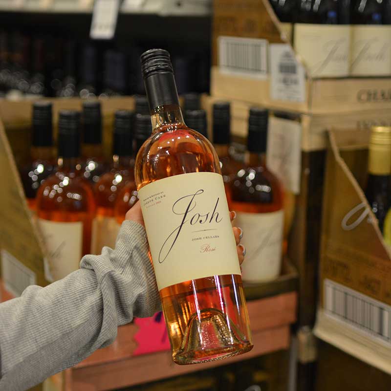 Hand holding a bottle of rosé wine in front of a wine selection at Hickey's Wine & Spirits.